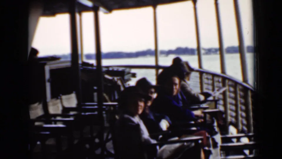 1940 Ontario home-movie frame of passengers in hats and coats seated on a covered boat deck with lake scenery beyond the windows.