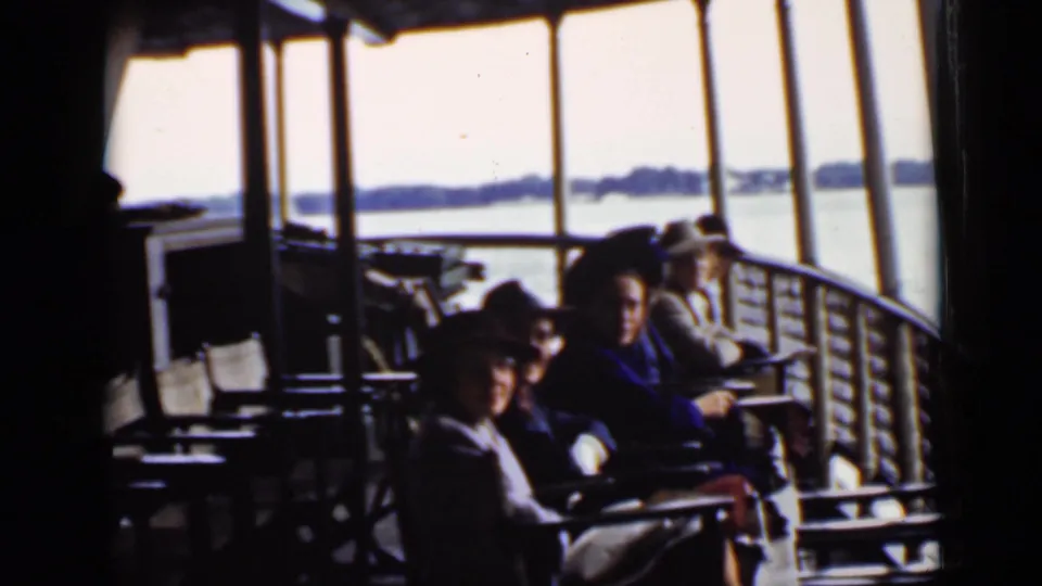 Passengers aboard an Ontario boat sit near deck railings and windows, with bright lake water and shoreline visible in the background.