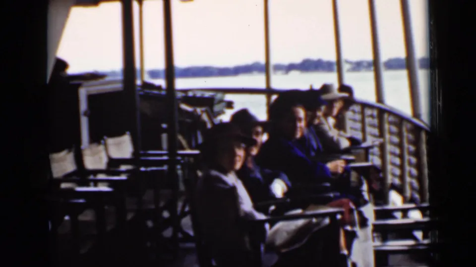 A passenger in a brimmed hat sits closest to the camera while others relax on a covered Ontario boat deck overlooking the lake.
