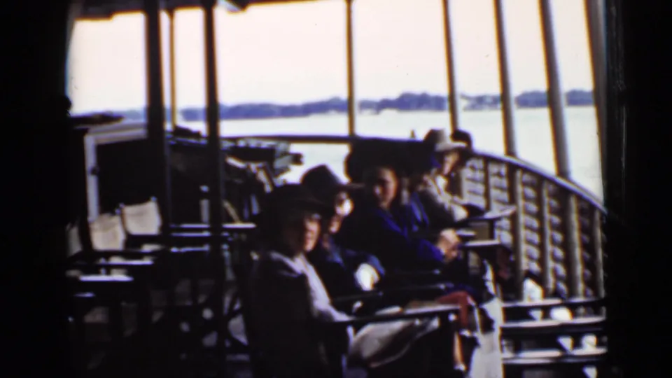 Ontario home-movie frame showing passengers seated under a covered boat deck, with railings, windows, bright water, and distant shoreline visible.