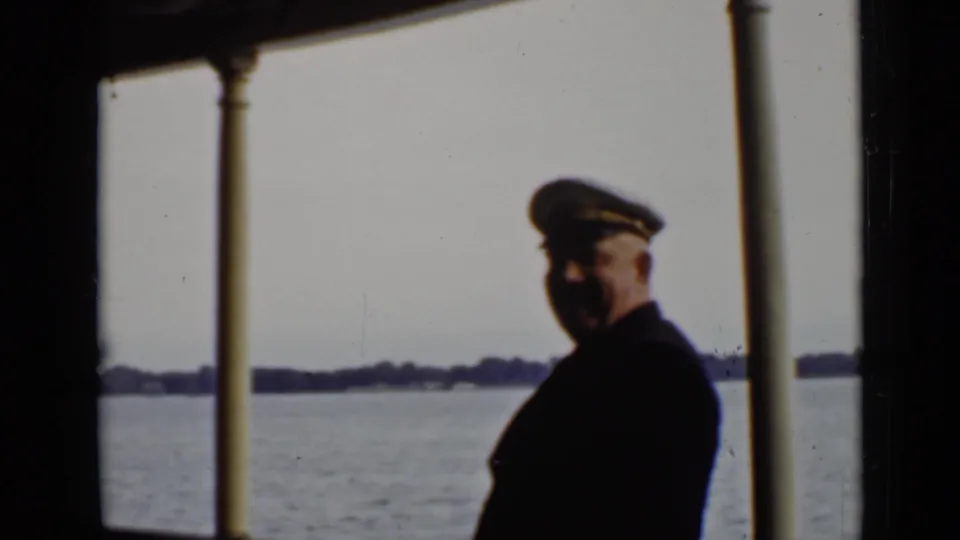 A uniformed ship officer or captain stands on the vessel deck in Ontario, with open water and shoreline behind him.