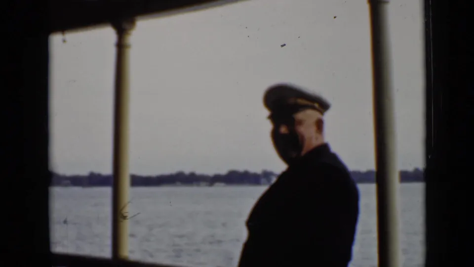 The uniformed man is seen in soft focus on the ship deck, framed by posts and a distant Ontario shoreline.