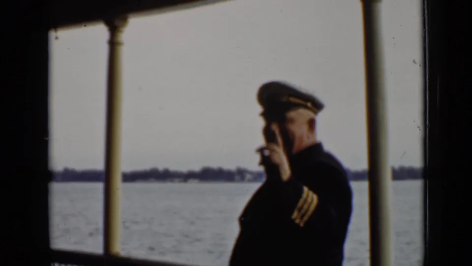 A uniformed ship officer or captain smiles and points toward the camera on a 1940 Ontario boat deck, with water behind him.