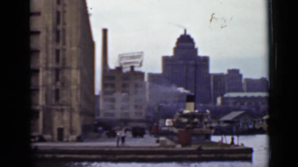 Ontario industrial waterfront with dock, smoke or steam, distant skyline, and small people visible near the quay.