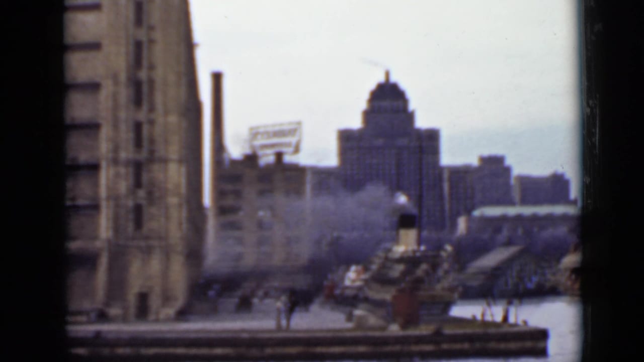 City harbor seen from a ferry window with smokestacks, piers, and a domed tower in the distance.
