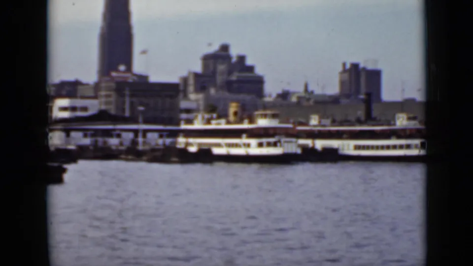 1940 Ontario waterfront frame showing rippled water, moored passenger boats, dockside buildings, and a tall skyline landmark.