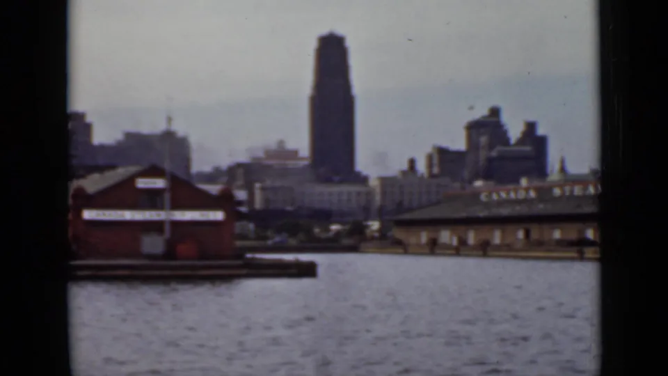 1940 Ontario waterfront scene with a tall tower, low dock buildings, and a partially visible sign on a roofline across the water.
