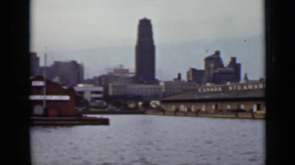 1940 Ontario harbor frame showing rippled water, low waterfront structures, a roofline sign, and a tall tower in the background.