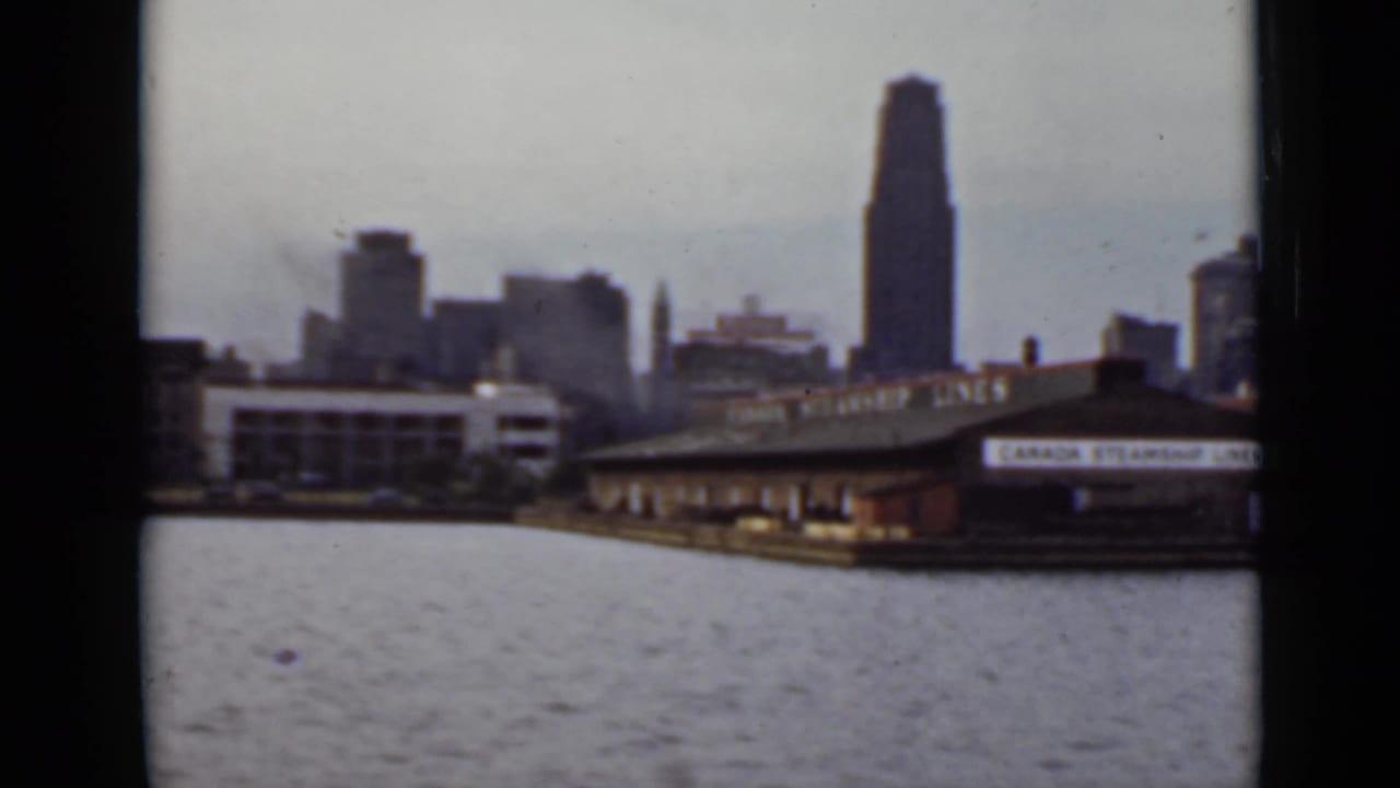 1940 Ontario waterfront view from a boat, with a tall skyscraper, river foreground, and Canada Steamship Lines terminal area.