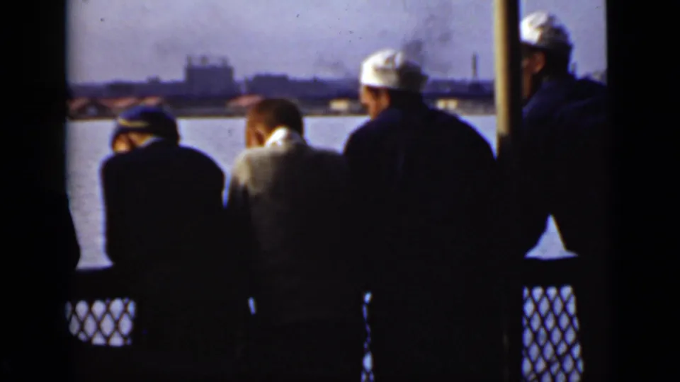 Several men, including sailors in white caps, stand at a boat railing in Ontario, looking toward an industrial shoreline.