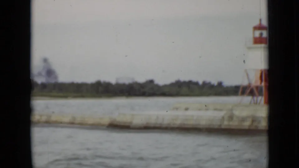 Ontario waterfront frame from 1940 with a red-and-white lighthouse or pier light on a concrete pier beyond choppy water.