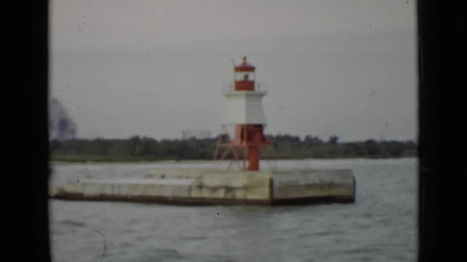 1940 Ontario harbor view from a boat, showing a red and white lighthouse on a concrete pier with trees beyond.