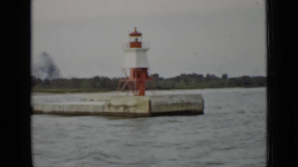 1940 Ontario boat-view frame of a small harbor lighthouse on a pier, with gray water and a low wooded shore behind it.