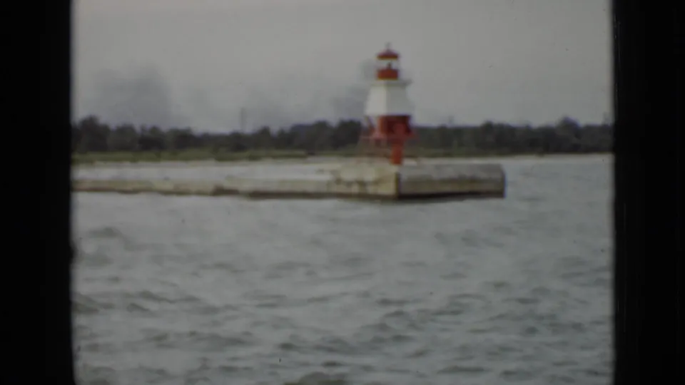 1940 Ontario waterfront frame with a small lighthouse on a concrete pier, seen across choppy water from a passing boat.