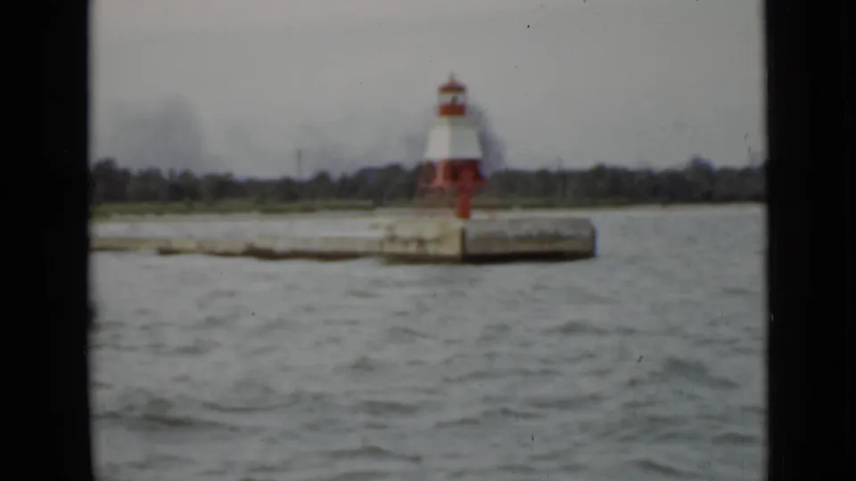 1940 Ontario 8mm frame showing a red-and-white lighthouse on a concrete pier, filmed from a moving boat across choppy gray water.