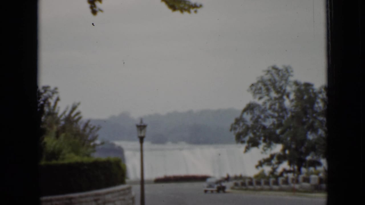 1940 Ontario home-movie view toward Niagara Falls with trees, a lamppost, riverside path, and parked car.