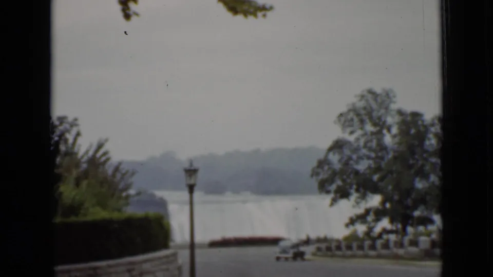 1940 Ontario home-movie view toward Niagara Falls with trees, a lamppost, riverside path, and parked car.