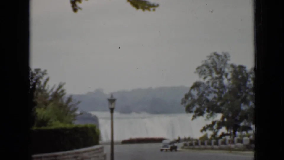 Niagara Falls in Ontario appears beyond trees and a lamppost, with a path and parked car in the foreground.