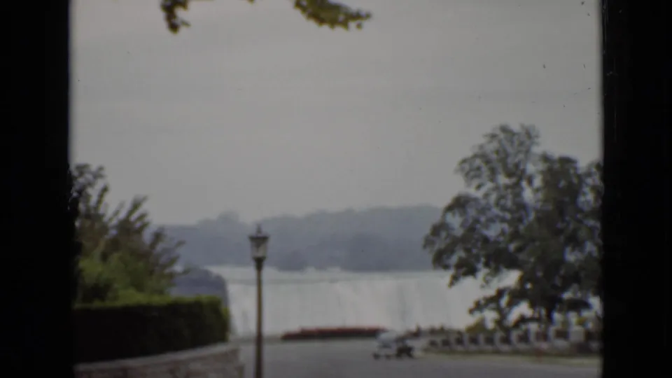 A soft 8mm view of Niagara Falls from Ontario, framed by trees, a lamppost, railings, path, and car.