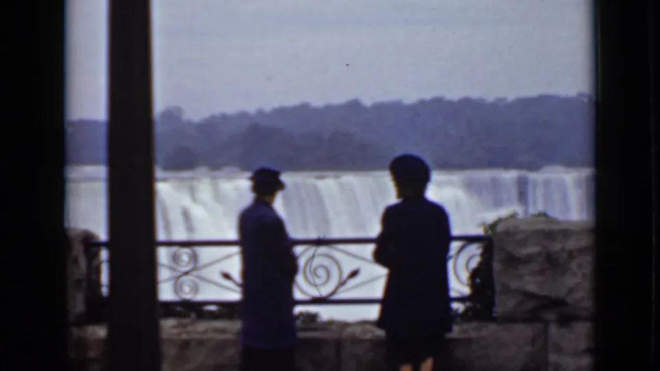 Two visitors in dark coats and hats stand on a Canadian-side Niagara overlook with American Falls and mist behind them.
