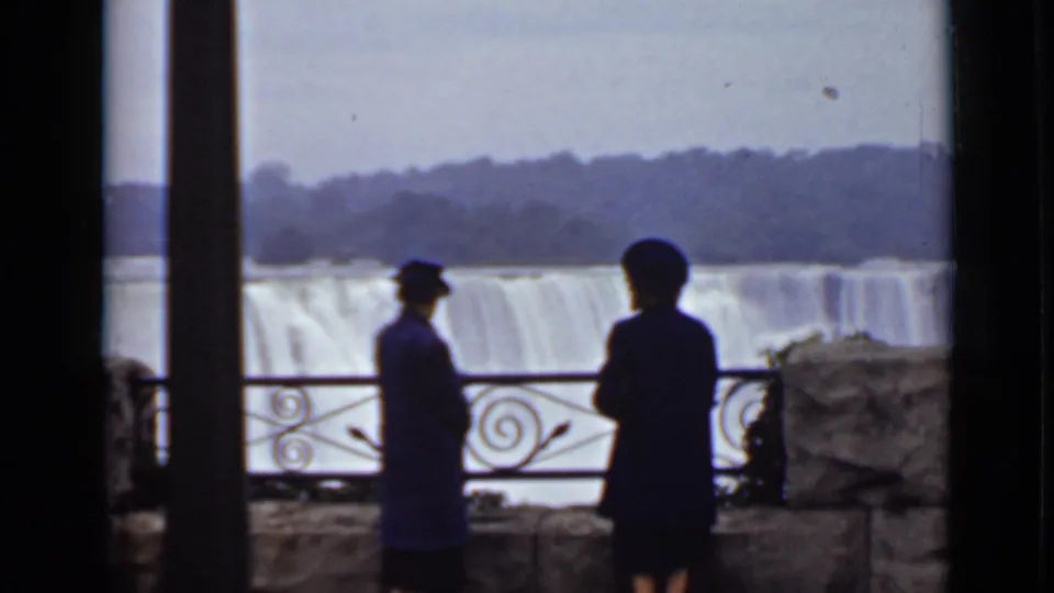 Two visitors in period coats stand near the decorative railing at Niagara Falls, with American Falls visible in soft focus beyond.