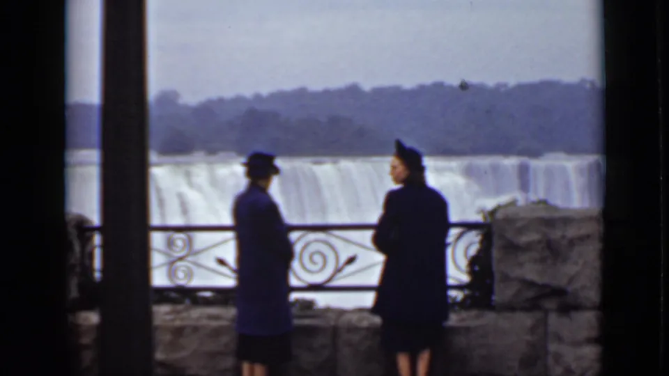At a Canadian-side Niagara overlook, two visitors in dark outerwear stand near the railing while American Falls fills the background.