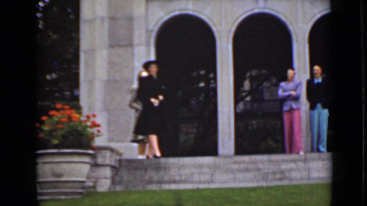In 1940s Ontario footage, several women in hats and suits stand outside a stone building with arched openings and red flowers nearby.