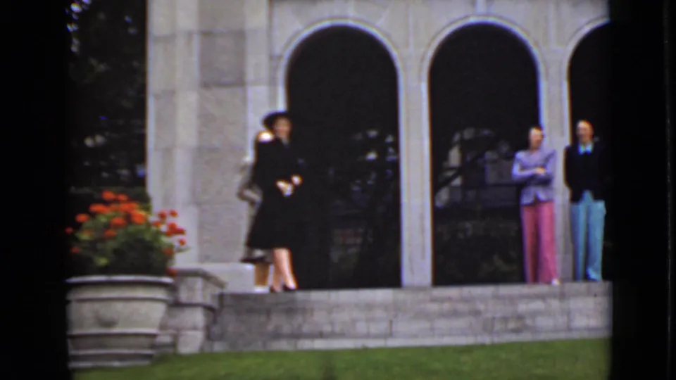 In 1940s Ontario footage, several women in hats and suits stand outside a stone building with arched openings and red flowers nearby.