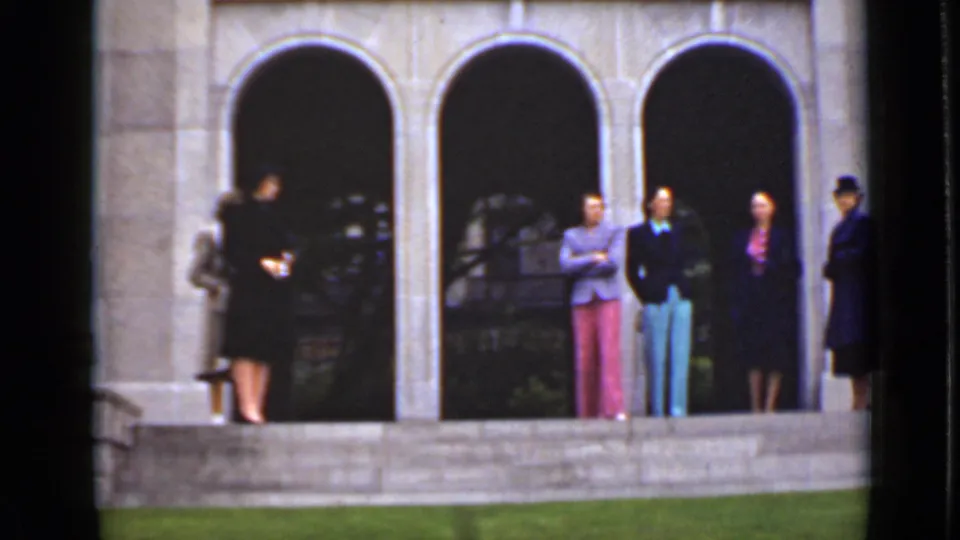 The 8mm frame shows women in hats, dresses, and bright suits waiting on stone steps outside an arched building in Ontario.