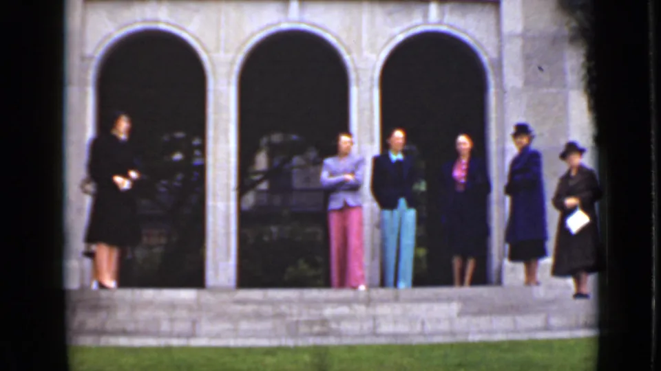 At the end of the shot, women stand across the stone steps, including one at right holding a pale paper or program.