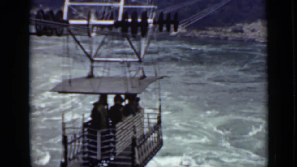 Vintage 8mm frame of a roofed cable gondola and overhead hardware crossing rough water in Ontario.