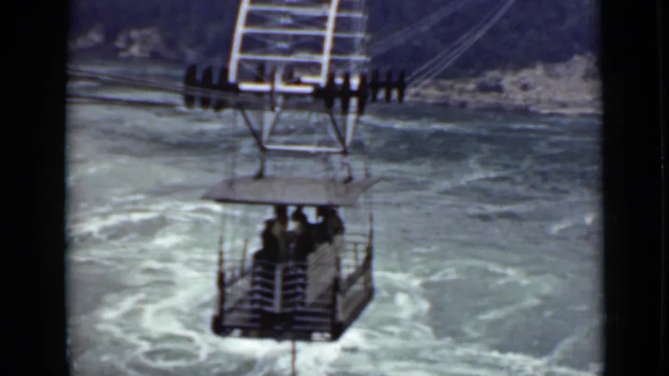 Open-air gondola in Ontario, 1940, carrying riders across a turbulent body of water beneath cable supports.