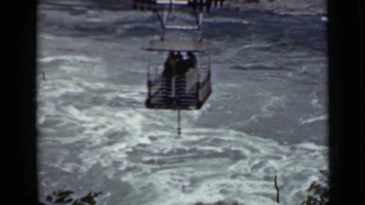 Open cable car hangs above rough river water with passengers visible inside.