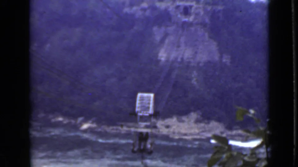 Faded 1940 Ontario home-movie frame showing the cable car over a river near the far bank, with foreground foliage.
