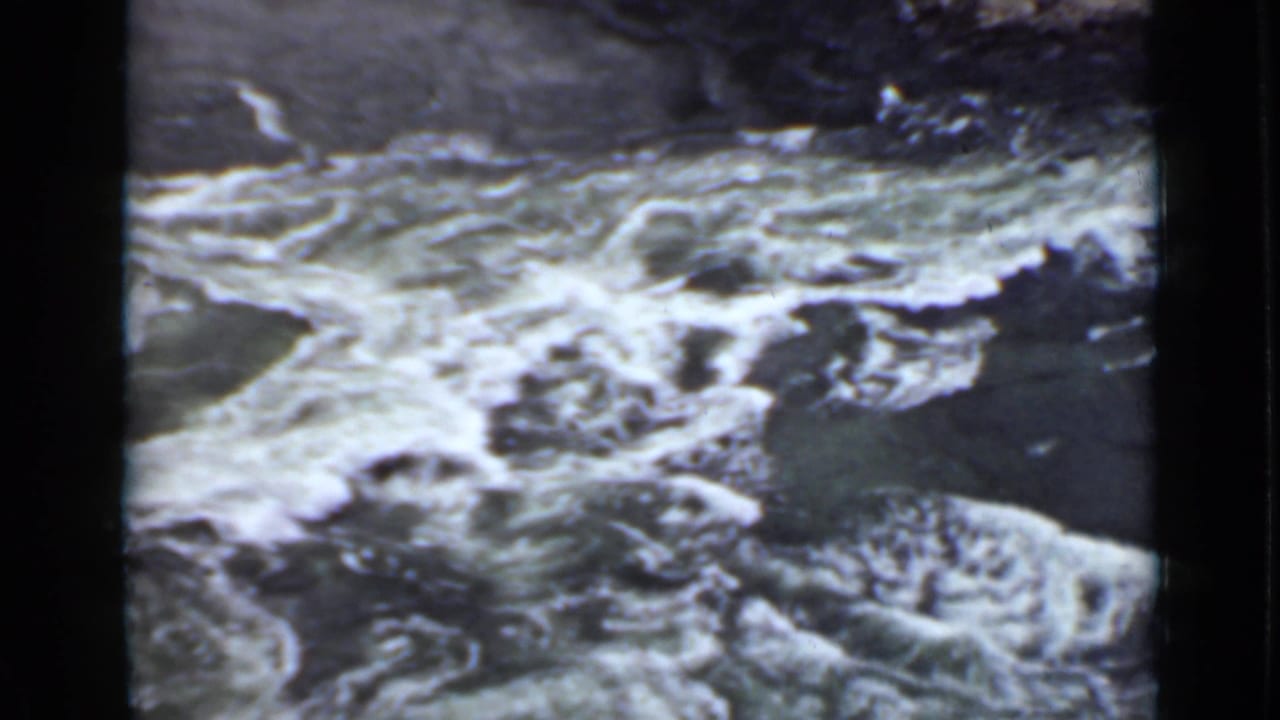 Whitewater river rushing around dark rocks below a shadowed bank.