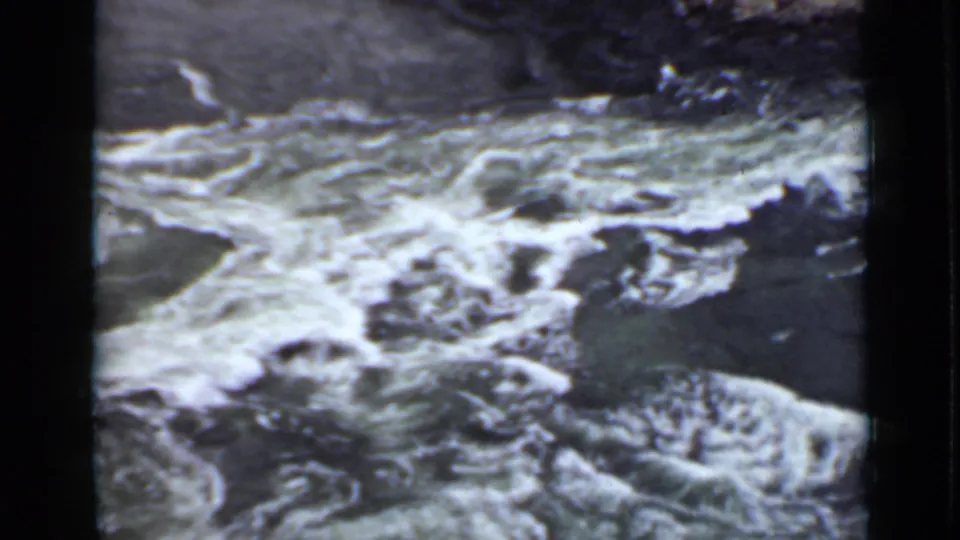 1940 Ontario 8mm frame showing turbulent river water breaking around rocks beneath a steep, shadowed shoreline.