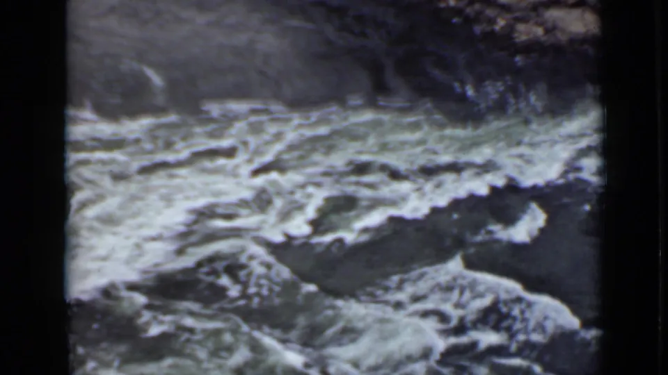 1940 Ontario home-movie frame with foaming rapids, exposed rocks, and a dark hillside or bank above the river.
