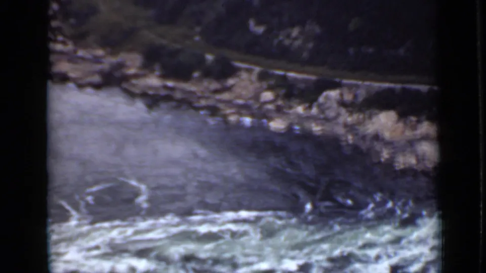 1940 Ontario home-movie frame of rapids beneath a curved rocky bank with a dark hillside rising behind it.