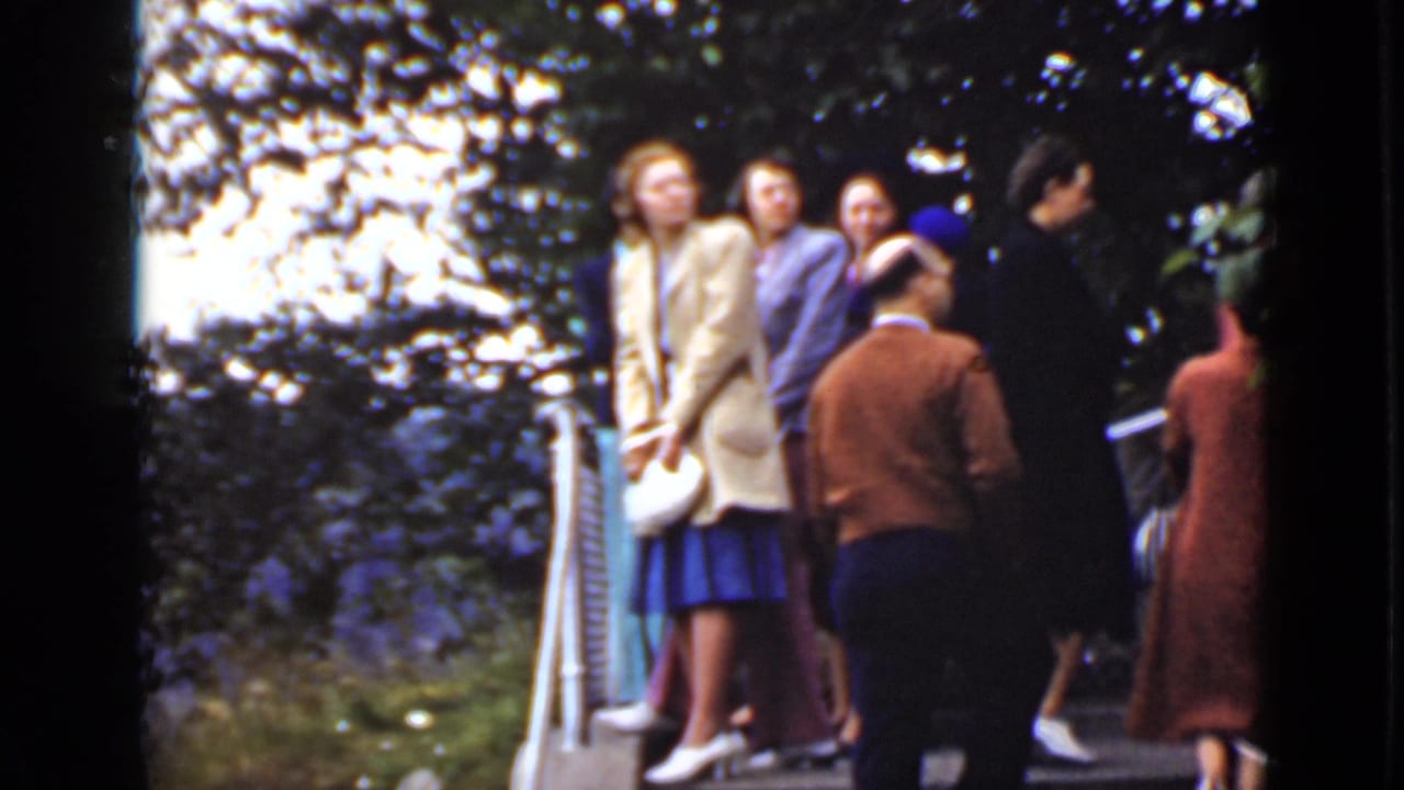 Adults in 1940s-style coats and hats gather on an outdoor deck near white railings.