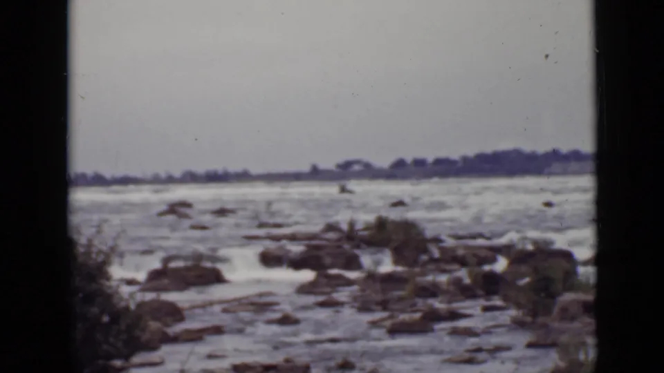 1940 Ontario shoreline view with churning water over scattered rocks and a low treeline in the distance.