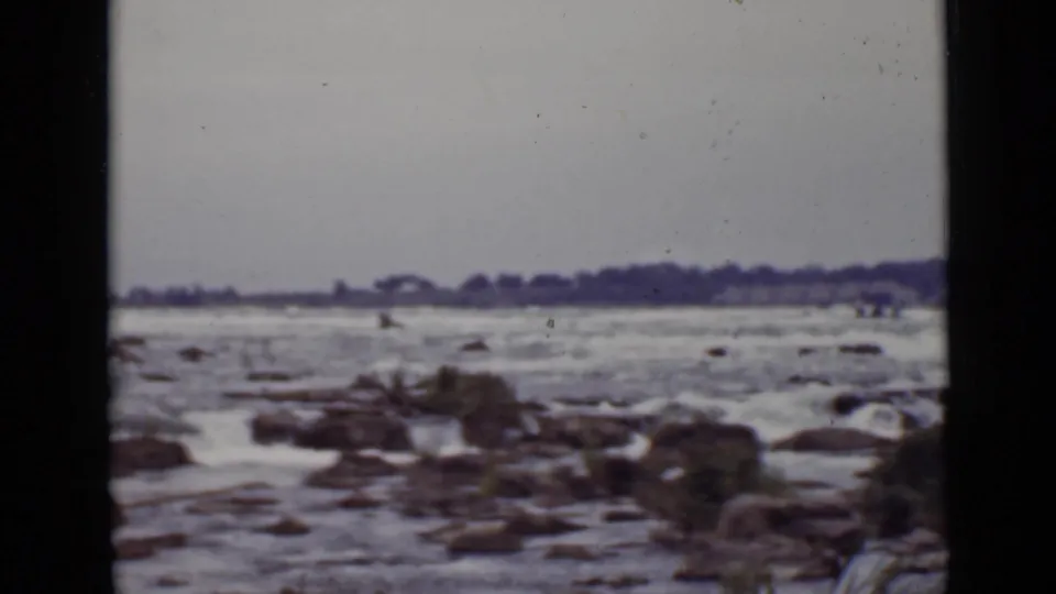 1940 Ontario home-movie view of turbulent water, exposed rocks, and a distant tree-lined shore on a cloudy day.