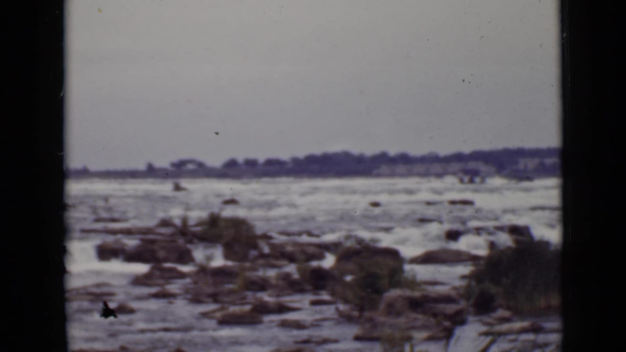 Fast water moving around rocks with a distant treeline under a pale sky.