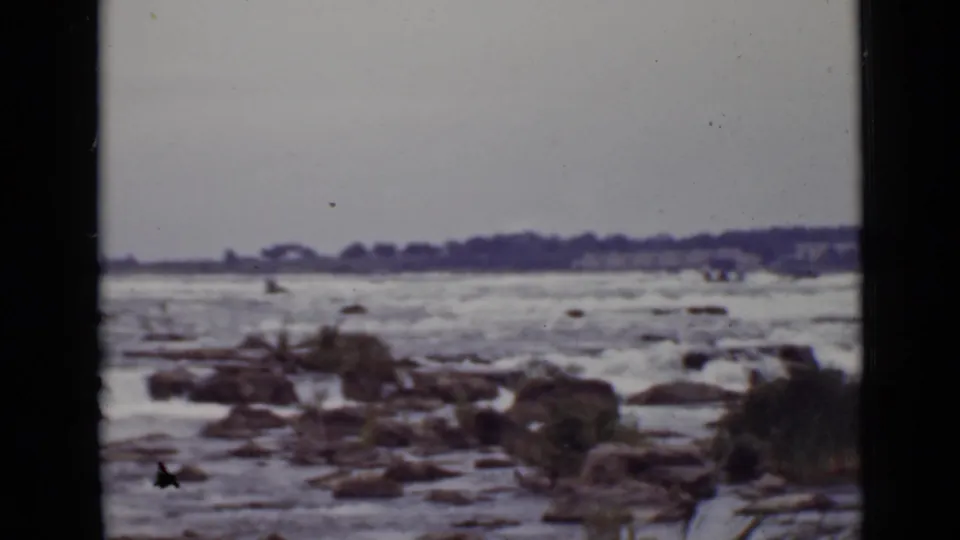 1940 Ontario 8mm frame showing rough river water, scattered rocks, low vegetation, and a distant treeline.