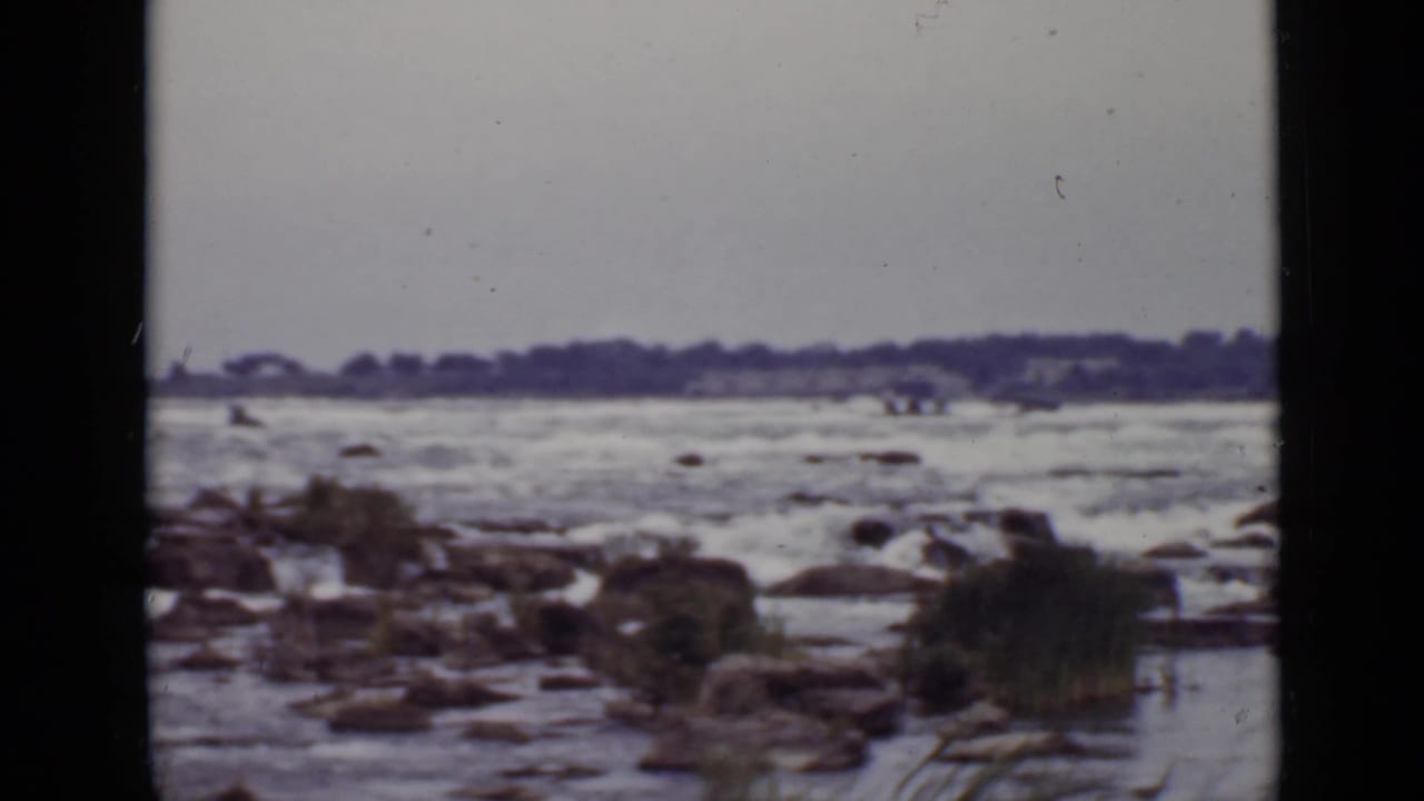 Rocky river shallows with churning water and a dark treeline in the distance.