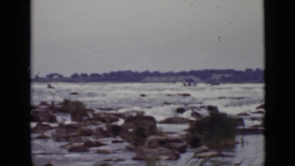 1940 Ontario home-movie frame of turbulent river water passing over rocks near a distant wooded bank.