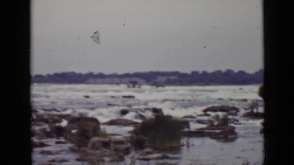 1940 Ontario 8mm view of fast-moving river water around rocks and reeds beneath an overcast sky.