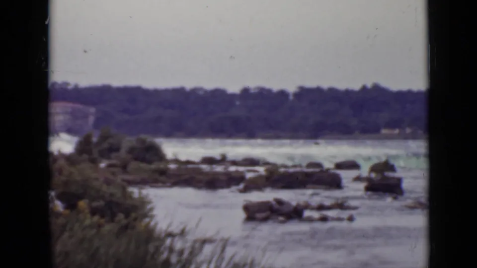 1940 Ontario home-movie frame showing rapids, rocks, shoreline reeds, and a wooded bank in the distance.