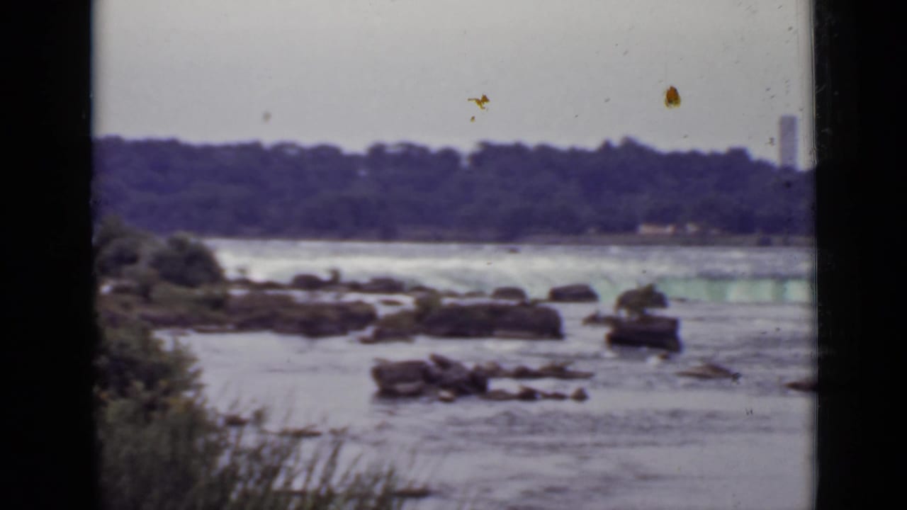Wide river view with rocks, rushing water, treeline, and a distant tower.