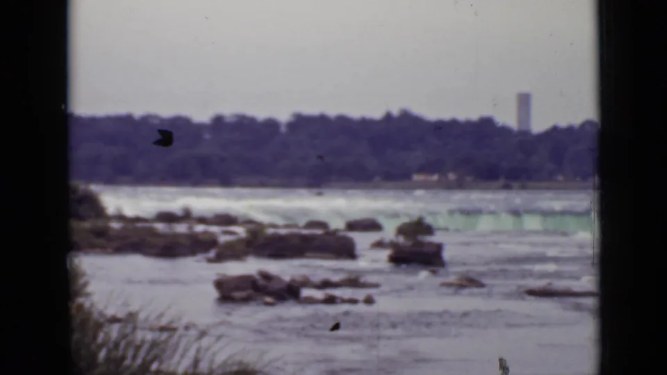 1940 Ontario home-movie frame of turbulent water and exposed rocks beneath a far treeline, with a narrow tower visible at right.