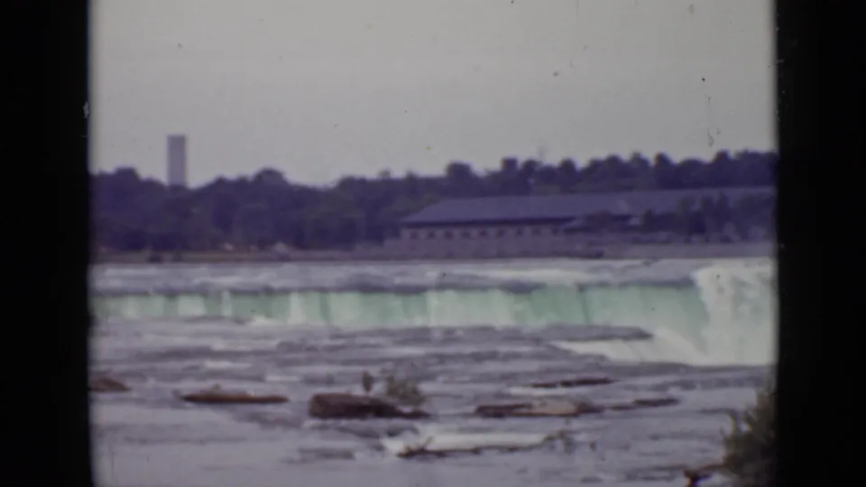 1940 Ontario 8mm view of a broad waterfall or dam edge, foreground rocks, far treeline, low buildings, and a distant tower.